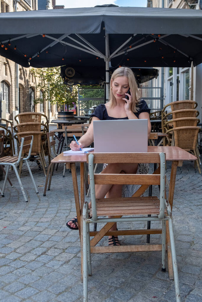 Akıllı telefonuyla laptopunu kullanan çekici sarışın iş kadını Avrupa 'da bir kentte tipik Belçika kafe terasında oturuyor. Yüksek kalite fotoğraf - Fotoğraf, Görsel