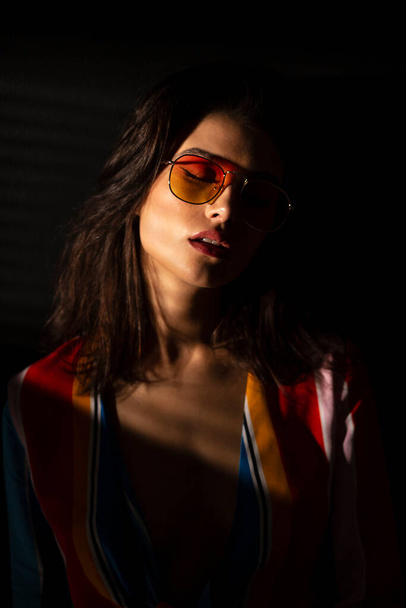 Closeup view of a young brunette woman in colorful clothes and sunglasses, black background. - Foto, Bild