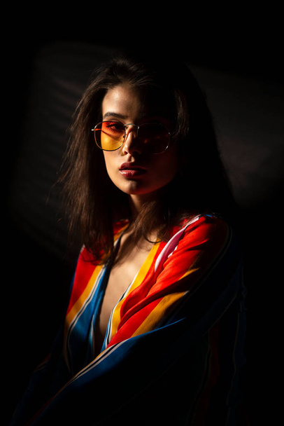Closeup image of a young brunette woman in colorful clothes and sunglasses, looking at camera, dark background. - Foto, Bild