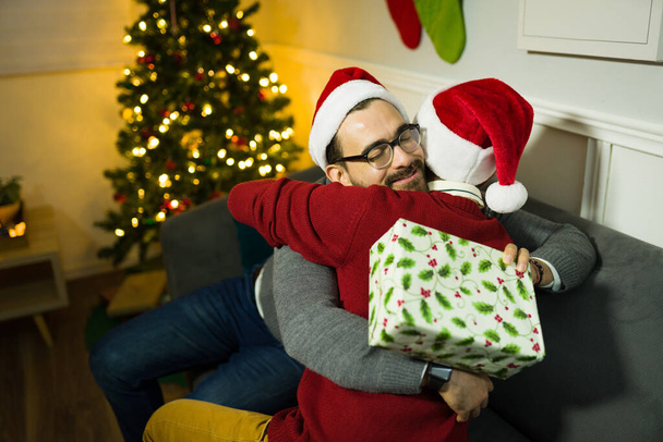 Rückansicht eines schwulen Paares, das sich umarmt und verliebt fühlt, während es zu Hause seine Weihnachtsgeschenke hält - Foto, Bild