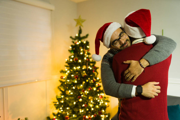 Ευτυχισμένος γκέι άντρας που αγκαλιάζει το σύντροφό του αφού έλαβε ένα δώρο μπροστά στο χριστουγεννιάτικο δέντρο του σπιτιού του. - Φωτογραφία, εικόνα