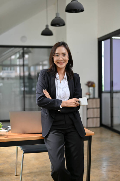 Retrato, mujer de negocios asiática segura y exitosa o gerente en traje formal apoyado en la mesa, brazos cruzados, sonriendo y mirando a la cámara. - Foto, imagen