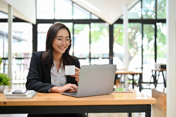 Exitosa y profesional millennial mujer de negocios asiática o gerente financiero femenino en gafas y traje formal a distancia trabajando en la cafetería, utilizando el ordenador portátil mientras disfruta de su café de la mañana. - Foto, imagen
