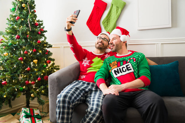 Pijama giyip Noel Baba şapkası takan, sosyal medya için selfie çeken mutlu ve çekici eşcinsel çift Noel 'i kutluyor. - Fotoğraf, Görsel