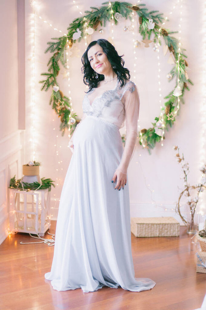 Young pregnant woman with dark hair in airy dress in a room decorated with pine needles and sparkling garlands for Christmas. Christmas mood . Pregnancy - Fotoğraf, Görsel