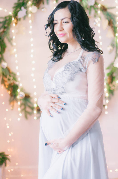 Young pregnant woman with dark hair in airy dress in a room decorated with pine needles and sparkling garlands for Christmas. Christmas mood . Pregnancy - Foto, imagen