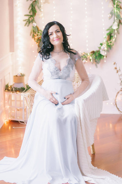 Young pregnant woman with dark hair in airy dress in a room decorated with pine needles and sparkling garlands for Christmas. Christmas mood . Pregnancy - Φωτογραφία, εικόνα