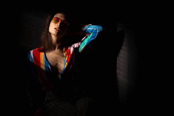 Portrait of a young brunette young woman in colorful clothes and orange sunglasses, with closed eyes, dark background. Horizontal view. - Photo, Image