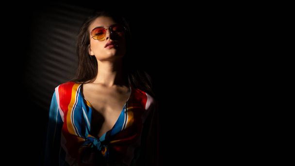 Portrait of a young brunette young woman in colorful clothes and orange sunglasses, with closed eyes, dark background. Horizontal view. Copy space. - Photo, Image