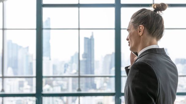 トップノットのヘアスタイルを持つ若いSMART CEOは、都市や都市の高層ビルの背景を持つ屋上テラスから彼の顧客を呼んでいます。町並みを背景に窓の隣に実業家が立つ - 写真・画像