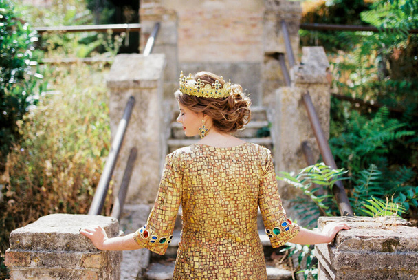 Νύφη με ένα χρυσό φόρεμα και στέμμα στέκεται στα πέτρινα σκαλοπάτια κρατώντας τους χειρολισθήρες. Υψηλής ποιότητας φωτογραφία - Φωτογραφία, εικόνα