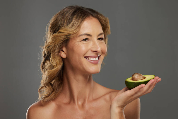Woman, happy and avocado in hand for health, wellness and detox care with face. Mature model, dermatology and smile with fruit diet for skincare, hair and beauty with natural facial cosmetic product. - Photo, Image