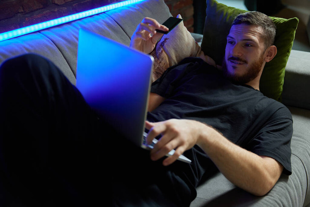 Cheerful handsome caucasian man lying on the sofa with laptop and credit card in hand in living room at home. Neon light. Online shopping and convenient payment method concept. Modern lifestyle - Foto, Imagem