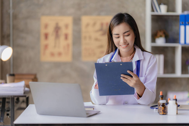 Azjatycki lekarz siedzący w recepcji szpitala, udzielający porad zdrowotnych i profilaktyki chorób, wygodne usługi online dla pacjentów i uśmiechnięty, wypisywanie recept na zamówienie leku. - Zdjęcie, obraz