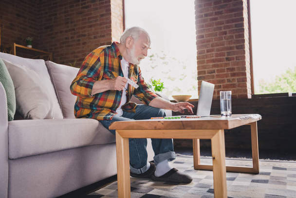Photo of depressed sad retired man dressed plaid shirt sitting couch reading pills prescription modern device indoors apartment. - Φωτογραφία, εικόνα