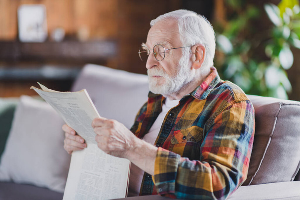 Photo of serious confident retired man dressed plaid shirt spectacles sitting couch reading fresh newspaper indoors apartment. - Φωτογραφία, εικόνα