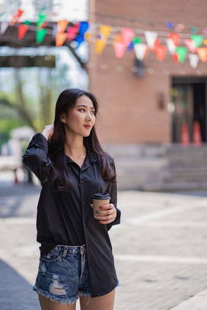 Uma mulher asiática bonita em uma camisa preta que prende uma xícara de café que está ao ar livre na frente de um shopping. - Foto, Imagem