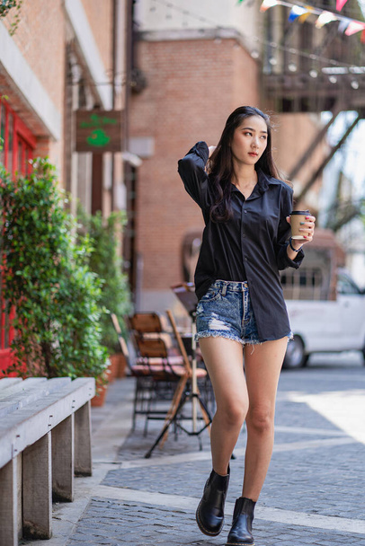 A pretty Asian woman in a black shirt holding a coffee cup standing outdoors in front of a shopping mall. - 写真・画像