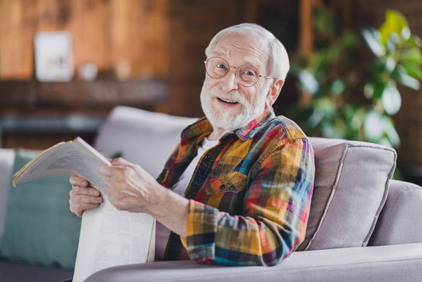 Photo of retired old man grandpa grey hair hold newspaper have break smile wear casual checkered clothes specs sit sofa indoors home room. - Fotó, kép