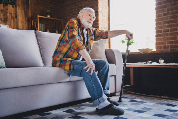 Фото стресованого нещасного чоловіка пенсіонер носить картату сорочку, що тримає прогулянкову тростину, намагаючись встати на диван у кімнаті будинку
. - Фото, зображення