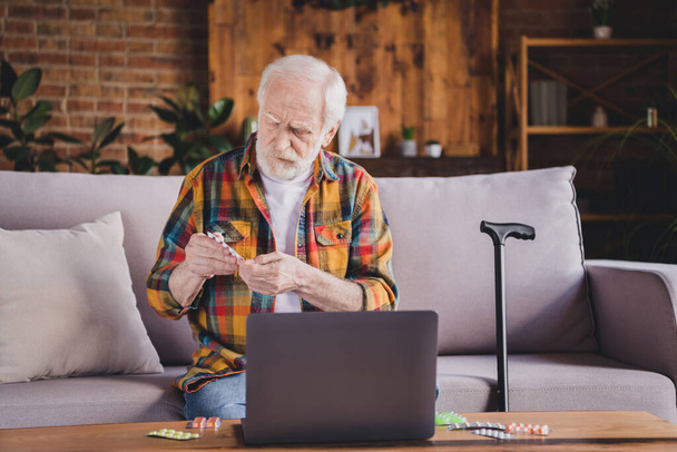 Photo of unhappy serious retired man dressed plaid shirt sitting couch reading pills dosage modern device indoors apartment. - Fotografie, Obrázek
