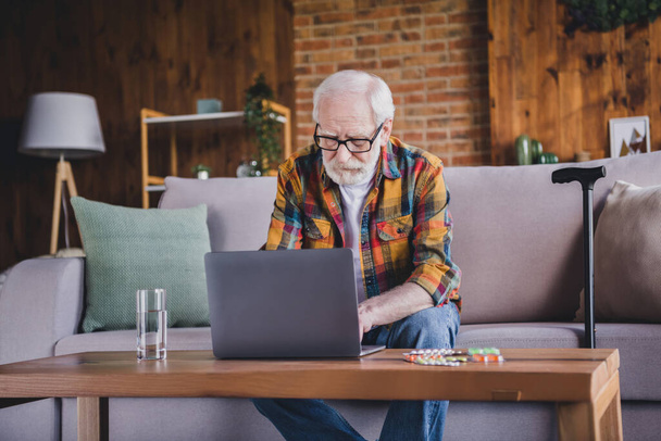 Photo of busy confident retired man dressed plaid shirt spectacles sitting couch reading pills dosage modern device indoors apartment. - Foto, afbeelding