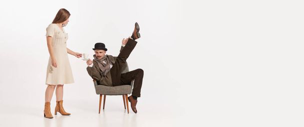 Porträt eines Mannes im Anzug, Mütze mit Schnurrbart auf einem Stuhl vor weißem Hintergrund sitzend. Frau schenkt ihm Kaffee ein. Konzept der Retro-Mode, Stil, Kreativität, Filmcharakter, Emotionen, Werbung. - Foto, Bild