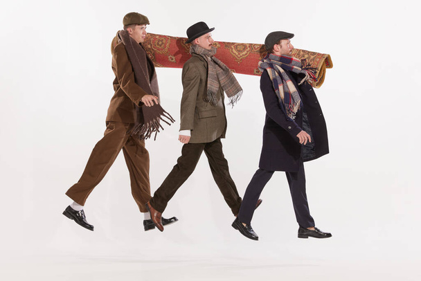 Portrait of three men in autumn clothes walking together and carrying carpet on shoulders over white background. Concept of retro fashion, style, friendship, creativity, youth culture, emotions, ad - Foto, Imagem
