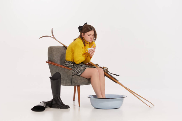 Portrait de jeune fille en pull jaune assis sur une chaise et réchauffant les jambes dans un bol avec de l'eau chaude. Après le ski. Concept de mode rétro, style, hiver, créativité, culture de la jeunesse, émotions - Photo, image