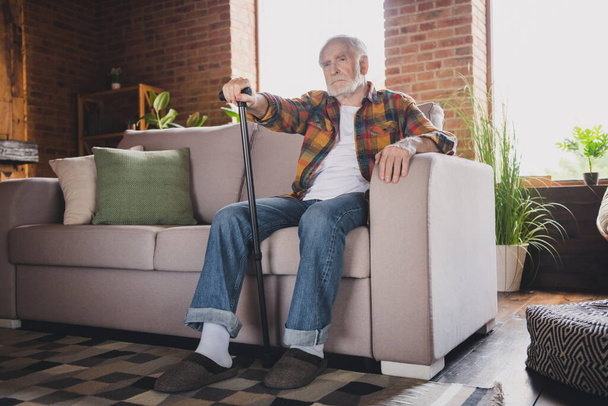 Photo of upset unhappy man pensioner wear checkered shirt sitting sofa holding walking cane indoors house room. - Foto, Imagen