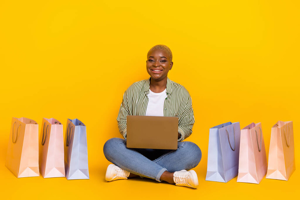 Full size photo of pretty positive lady sit floor use netbook shopping packages isolated on yellow color background. - Zdjęcie, obraz