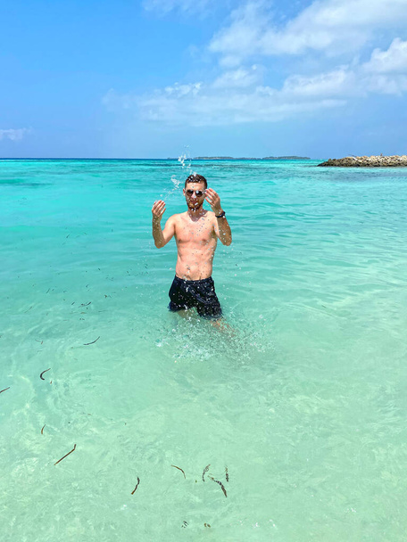 Handsome young man standing on the beach on an island in the Maldives, shirtless in boxer shorts, showing off his muscular body. Male standing in the ocean, splashing around in the water. - Photo, Image