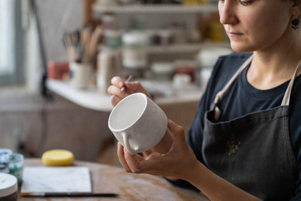 Craftswoman creates new masterpiece in work studio making sketch of future pattern on white mug. Lady master wants to decorate kitchen with painted clay dish using pencil to draw interesting pattern - Photo, image