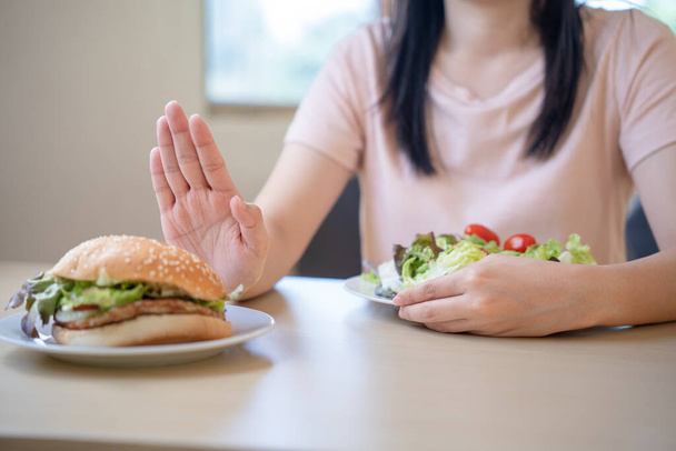 Healthy asian woman pushes a burger plate and choose to eat salads. Refuse fat and snacks with trans fats according to a diet plan and clean food. - Foto, afbeelding