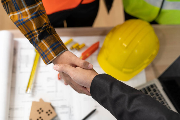 不動産屋は建設チームのリーダーと握手をする。ビジネスは、エンジニアがモデル構築プロジェクトを示した後にホームビルダーに署名することに同意します。住宅ローン、賃貸、購入、販売、購入、移動、販売の家. - 写真・画像