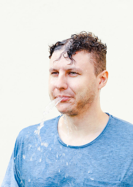 Natte jonge blanke man in blauw t-shirt met krullend haar spuugt water uit zijn mond op een witte achtergrond. Portret, regen, verticale foto. - Foto, afbeelding