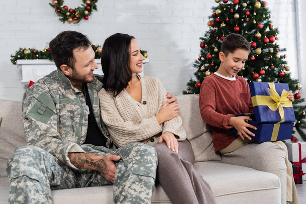 smiling boy opening gift box near mother and father in camouflage sitting on couch - Foto, Imagen