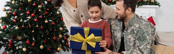 amazed boy opening christmas present near mother and father in camouflage, banner - Photo, Image
