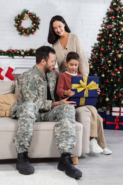 cheerful woman looking at son opening gift box near father in camouflage and decorated christmas tree - Photo, Image