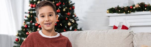 joyful boy in teeth brackets smiling at camera near christmas tree on blurred background, banner - Photo, Image