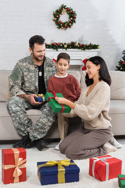 счастливая женщина сидит на полу с подарочной коробкой рядом сын и муж в камуфляже - Фото, изображение
