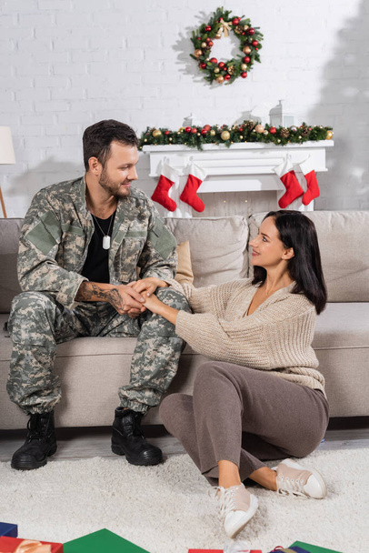 smiling woman sitting on floor and holding hands of husband in camouflage in living room with christmas decor - Photo, Image