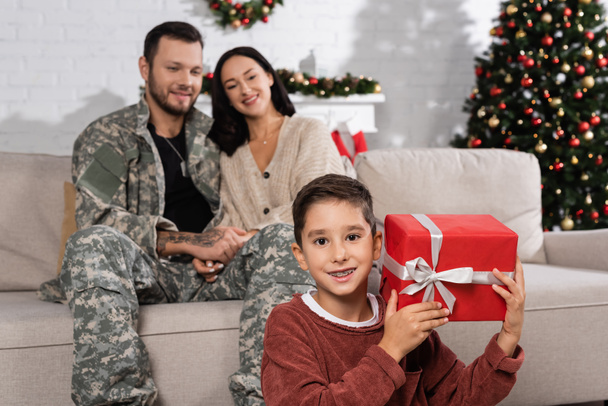 Junge mit Weihnachtsgeschenk lächelt in die Kamera neben Mutter und Vater in Tarnkleidung  - Foto, Bild