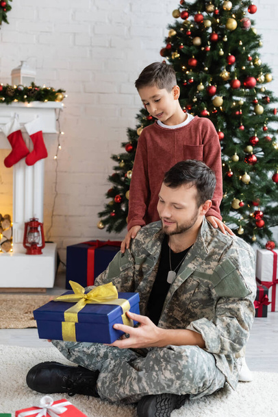 happy military man holding new year present near son and blurred christmas tree - Foto, Imagem