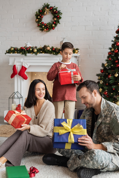 στρατιωτικό άνοιγμα κουτί δώρου άνθρωπος κοντά χαμογελαστός σύζυγος και γιος στο σαλόνι με χριστουγεννιάτικη διακόσμηση - Φωτογραφία, εικόνα