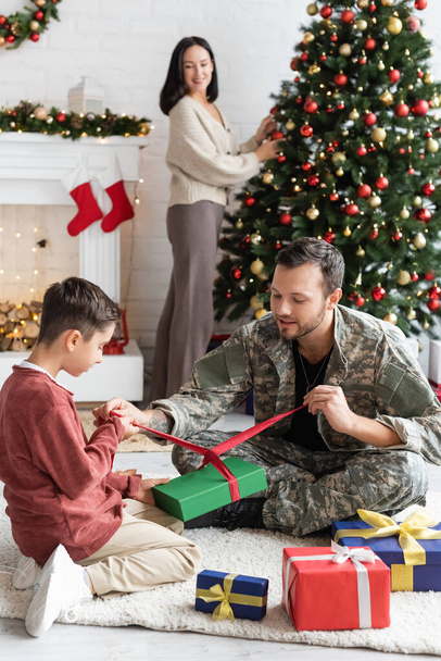 militar hombre embalaje regalo cajas con hijo cerca esposa decoración navidad árbol sobre fondo borroso - Foto, Imagen