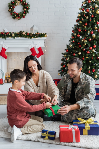 мужчина в камуфляже готовит подарки с женой и сыном возле рождественской елки - Фото, изображение