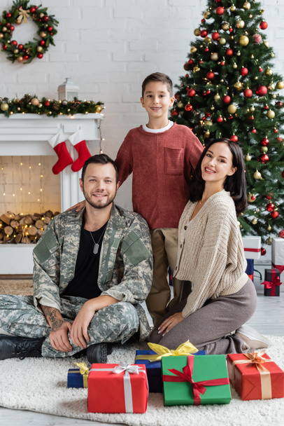 szczęśliwy wojskowy człowiek z żoną i synem patrząc na aparat w pobliżu pudełek i ozdobione choinki - Zdjęcie, obraz