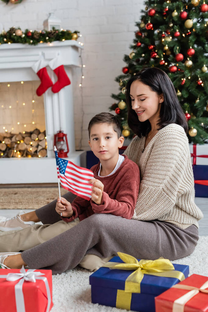 πατριωτικό αγόρι κρατώντας σημαία ΗΠΑ, ενώ κάθεται στο πάτωμα με χαμογελαστή μαμά κοντά χριστουγεννιάτικα δώρα - Φωτογραφία, εικόνα