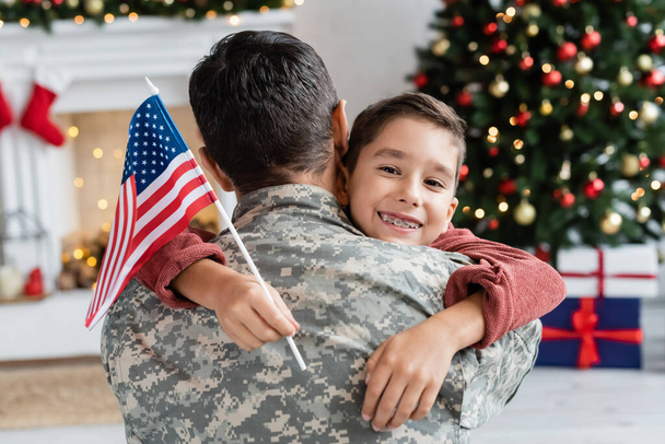 Glücklicher Junge in Zahnspange hält US-Fahne und umarmt Vater in Tarnkleidung - Foto, Bild